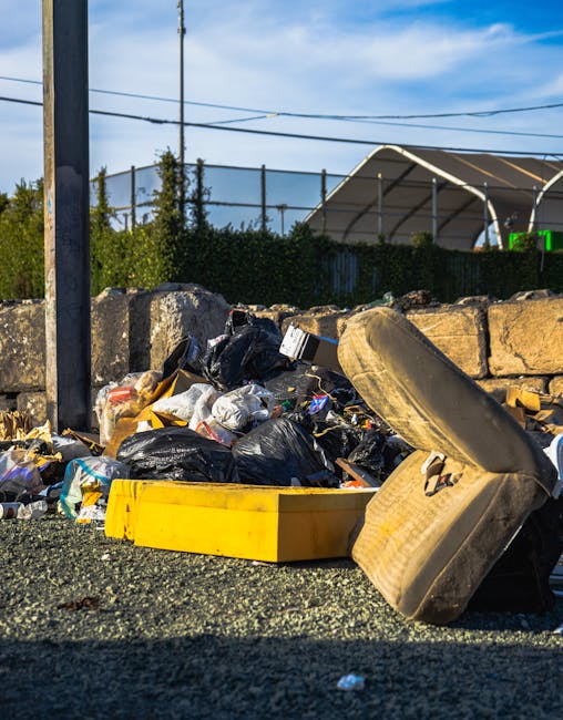 A pile of mixed domestic rubbish, including discarded black plastic bags, a yellow plastic storage bin, and an old, dirty car tire, all resting on a gravel surface in an outdoor setting. Behind the rubbish, there is a low stone wall made of rough, irregular concrete blocks, with green foliage and bushes growing along its top. In the background, a chain-link fence topped with barbed wire runs horizontally across the scene, partially obscuring a large, semi-circular fabric-covered structure with a metal frame, possibly an industrial or storage building. To the left, a metal pole supports overhead electrical wires that stretch across the sky, which is partly cloudy with patches of blue visible. The scene is illuminated by natural daylight, emphasizing the textures of the trash and surrounding environment, highlighting the informal or private waste collection context, typical of site clearance or alternative disposal methods outside standard municipal rubbish collection.