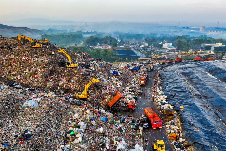 An expansive landfill site filled with a massive mound of mixed waste, including plastic bags, cardboard, and various refuse materials, dominates the foreground. Several yellow excavators are actively working on the refuse heap, scooping and moving the trash, their metal surfaces reflecting ambient light. To the right, a narrow, winding access road runs alongside the landfill, with an array of trucks in red, orange, and green actively engaged in transporting waste debris. Some trucks are partially loaded, with refuse spilling from their open tops, while others are waiting for their turn to unload or move along the site. The ground surrounding the road appears muddy and uneven, with scattered smaller waste items and debris. In the background, low-lying buildings and industrial structures are visible, set against a hazy, overcast sky, hinting at a busy waste disposal operation. The overall scene reflects a large-scale waste management process, possibly managed by independent contractors like Waste Disposal Enfield, emphasizing the scale and activity involved in extensive rubbish handling and disposal tasks under an overcast, dim light atmosphere.