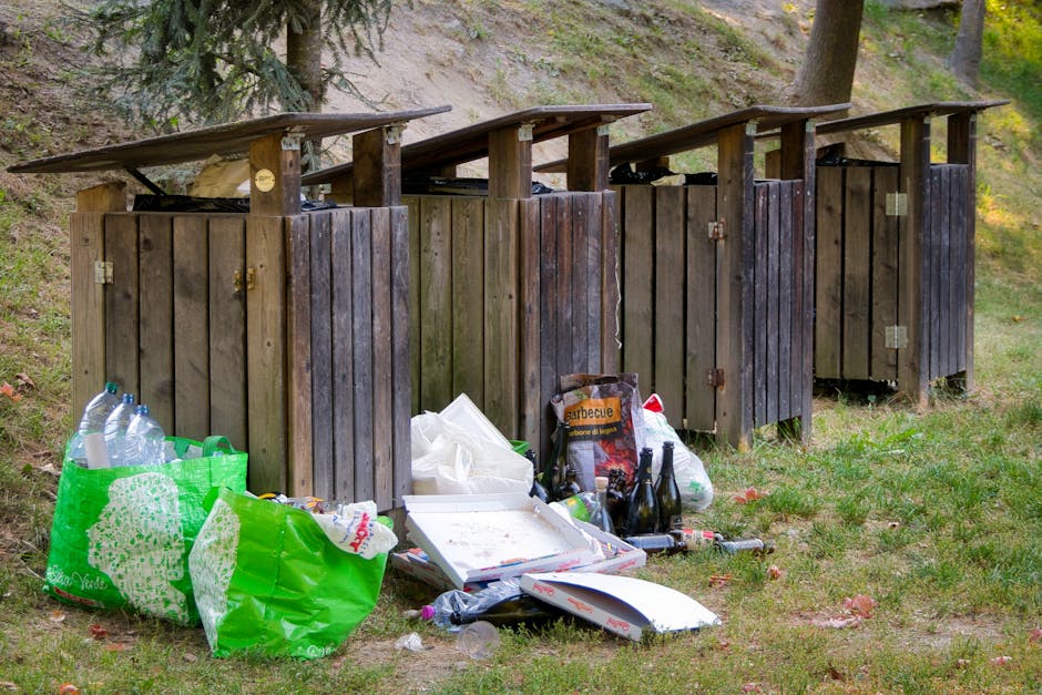 Four wooden outdoor rubbish bins with hinged lids are aligned on a grassy area beside a slope with trees and bushes in the background. The bins are weathered and dark brown, with some showing signs of wear and slight warping. In front of the first bin, there are several large green plastic shopping bags filled with plastic bottles and other discarded items, along with a white pizza box, a paper bag labelled 'barbecue', and a few empty glass bottles scattered on the grass. The scene appears outdoors in a natural environment, possibly within a park or a private garden, with daylight illuminating the scene. The presence of discarded waste indicates improper disposal, contrasting with professional rubbish removal services such as those offered by Waste Disposal Enfield, which typically handle waste collection in a clean and organized manner to prevent environmental impact.