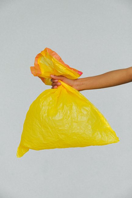 A person's arm extending from the right side of the image holding a yellow plastic rubbish bag with a crinkled texture, torn open at the top to reveal orange and yellow waste material inside. The bag is filled, creating a rounded shape, and is held against a plain, light-colored background. The scene suggests an act of rubbish collection or disposal, supported by the context of waste management services. The lighting is even, emphasizing the glossy surface of the plastic bag, which is typical for waste collection activities. This image visually relates to on-site refuse handling or independent waste disposal tasks, such as rubbish removal performed outside a property or during waste clearance operations, in line with services offered by Waste Disposal Enfield and similar providers.