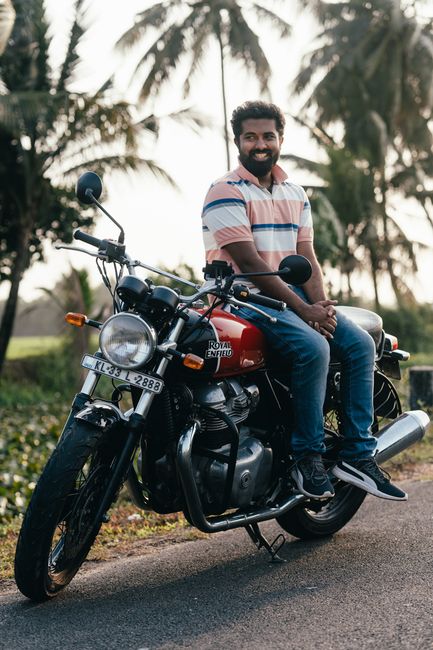 A young man with a dark beard and curly hair is sitting on a classic red and black Royal Enfield motorcycle parked on a paved road. He is wearing a short-sleeved, striped polo shirt in shades of pink, blue, and white, along with blue jeans and dark sneakers. The motorcycle features a prominent round headlight, black handlebars with rearview mirrors, and a visible license plate reading 'KL 35 L 2888.' The background consists of tall coconut trees and lush green foliage, indicating a tropical or rural environment, with soft, natural daylight illuminating the scene. The setting suggests a relaxed outdoor environment, possibly during a warm day, with the motorcycle and the individual as focal points, aligning with private transportation or lifestyle imagery. The image captures a casual, everyday moment in an outdoor space, suitable for illustrating themes of commuting, outdoor leisure, or independent travel related to waste disposal services such as waste management or rubbish removal, although no specific equipment or waste objects are visible in this context.