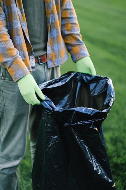 A person dressed in a checkered yellow and grey jacket, green disposable gloves, and grey trousers is holding open the top of a large black plastic rubbish bag with both hands. The bag is partially open, revealing an empty interior, and is positioned outdoors on a grassy area. The background features a well-maintained lawn with lush green grass, suggesting a garden or park environment. The scene appears to depict a waste collection or rubbish removal activity, possibly indicating a garden clearance or outdoor cleaning task, which aligns with private waste disposal services offered by Waste Disposal Enfield. The lighting is natural, with soft ambient daylight illuminating the scene, emphasizing the textures of the plastic bag’s glossy finish and the fabric of the individual’s clothing.
